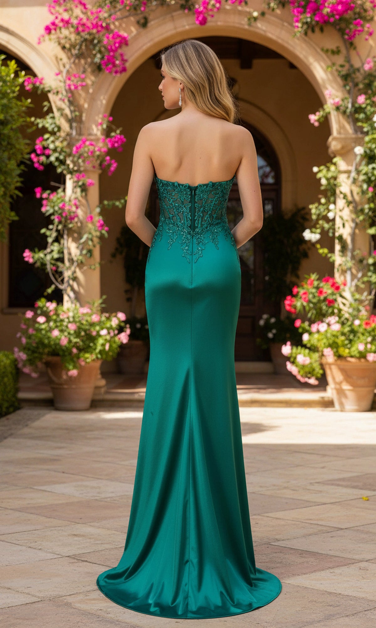 A woman wearing the PromGirl Exclusive 250247, a strapless emerald green prom dress with lace embroidery and a sheer corset bodice, stands outdoors among potted plants and blooming arches.
